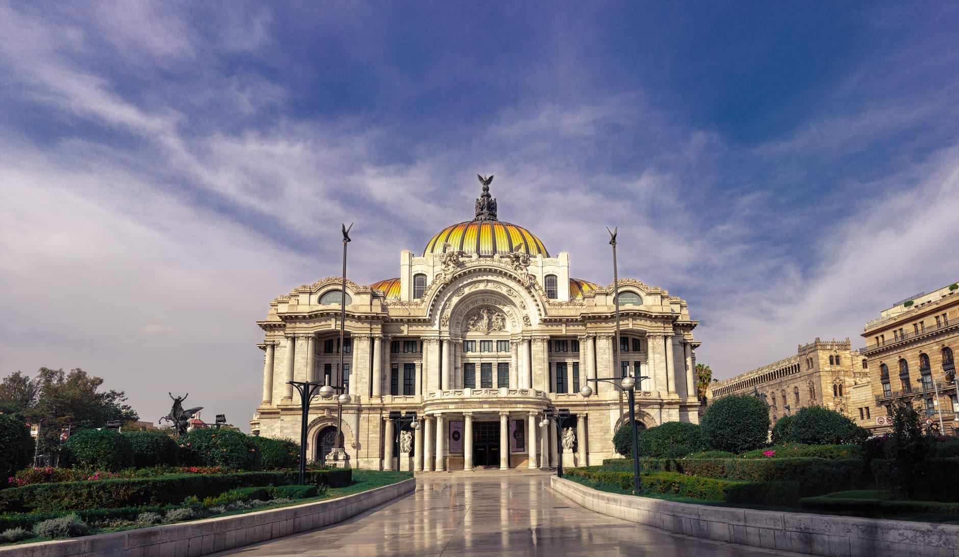 Vista frontal do Palacio de Bellas Artes, o icônico marco de mármore e cúpula de vidro no Centro Histórico da Cidade do México, com céu azul e jardins ornamentais.