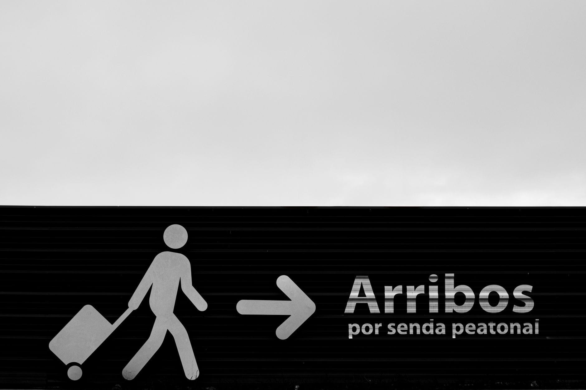 Placa em preto e branco em um aeroporto mostrando um viajante com mala e a palavra 'Arribos' para chegadas.