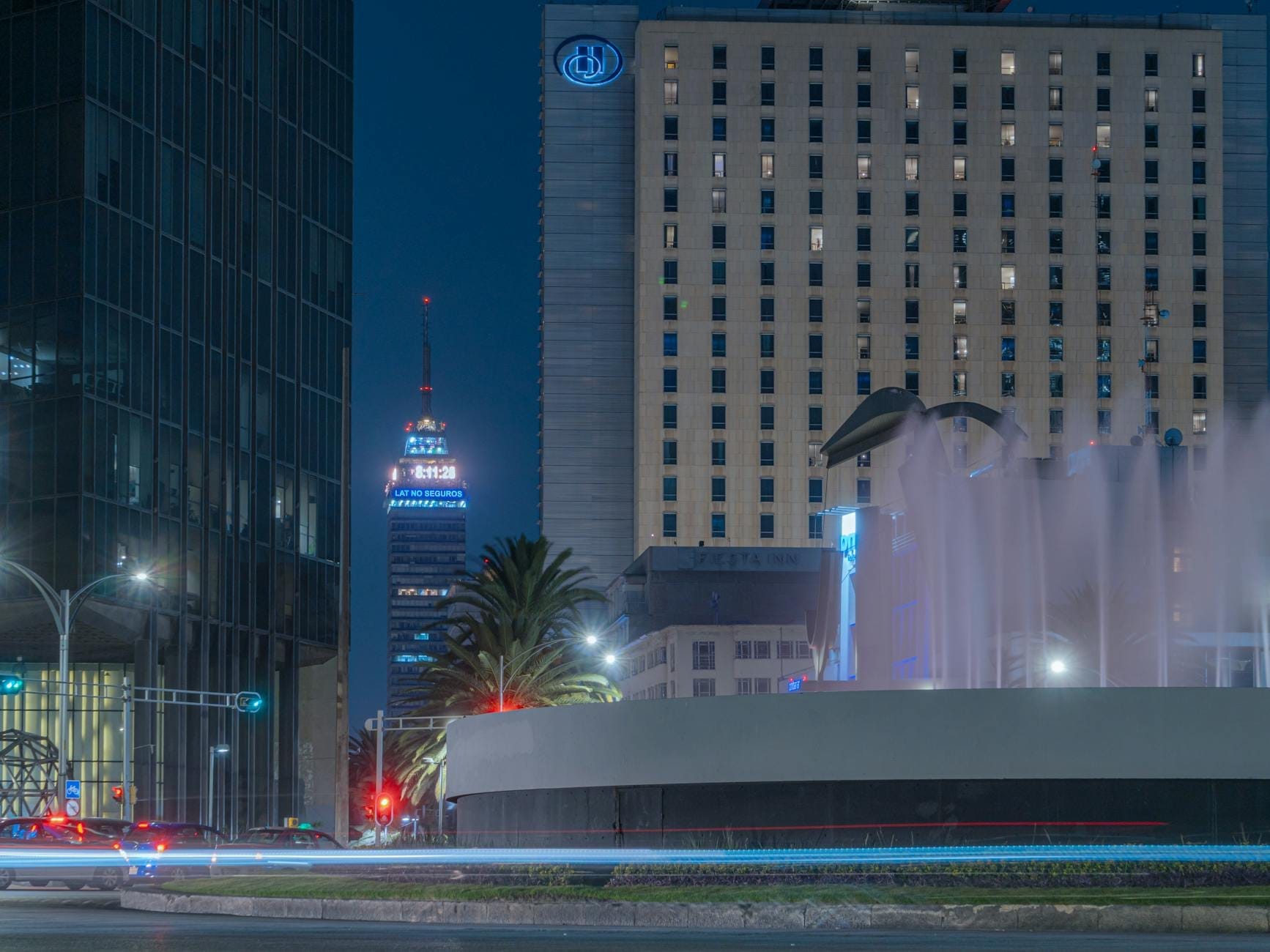 Vista noturna de hotéis de luxo e edifícios comerciais com janelas iluminadas e uma fonte no centro da Cidade do México, evocando uma atmosfera urbana sofisticada.