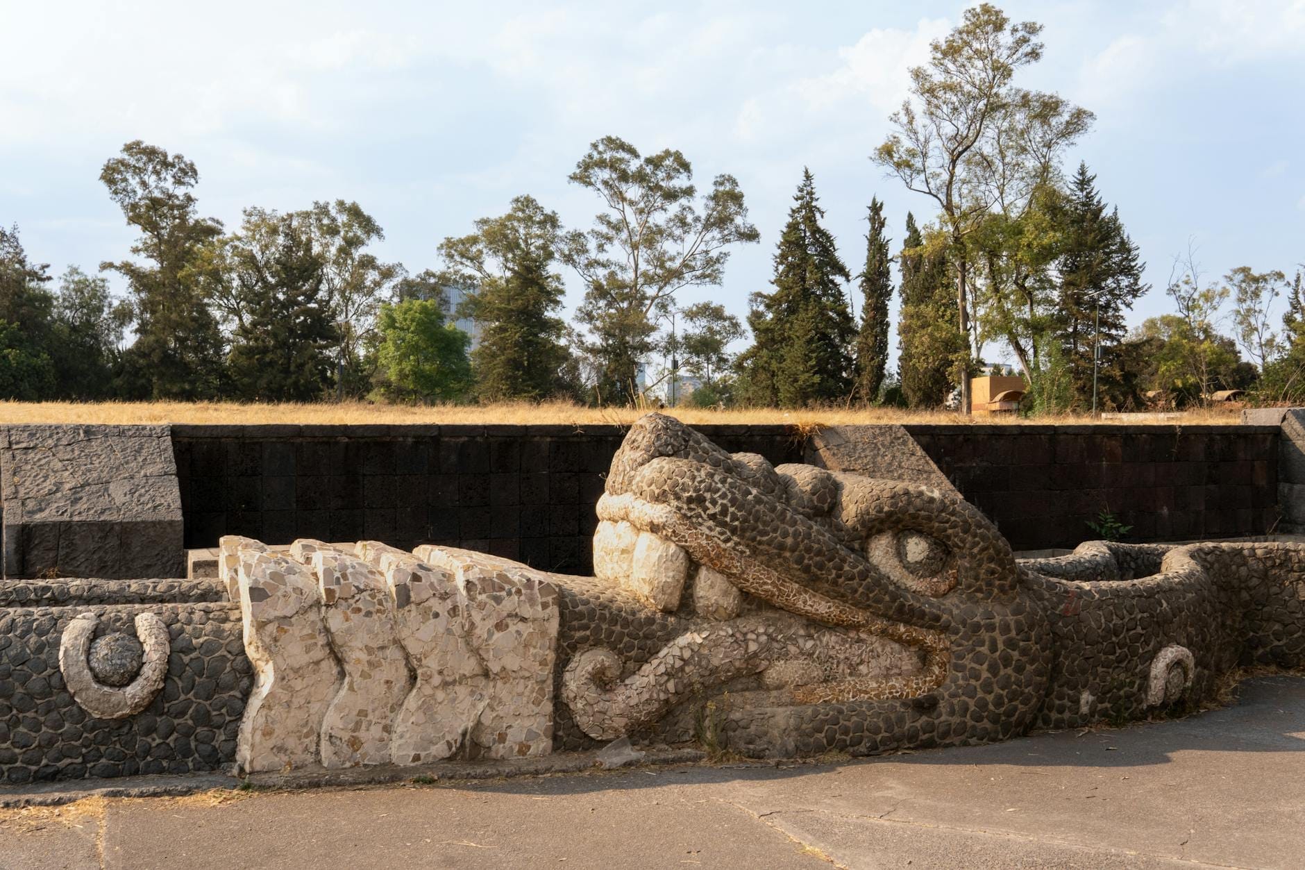 Cabeça de serpente esculpida em pedra em um sítio arqueológico ao ar livre cercado de árvores na Cidade do México.