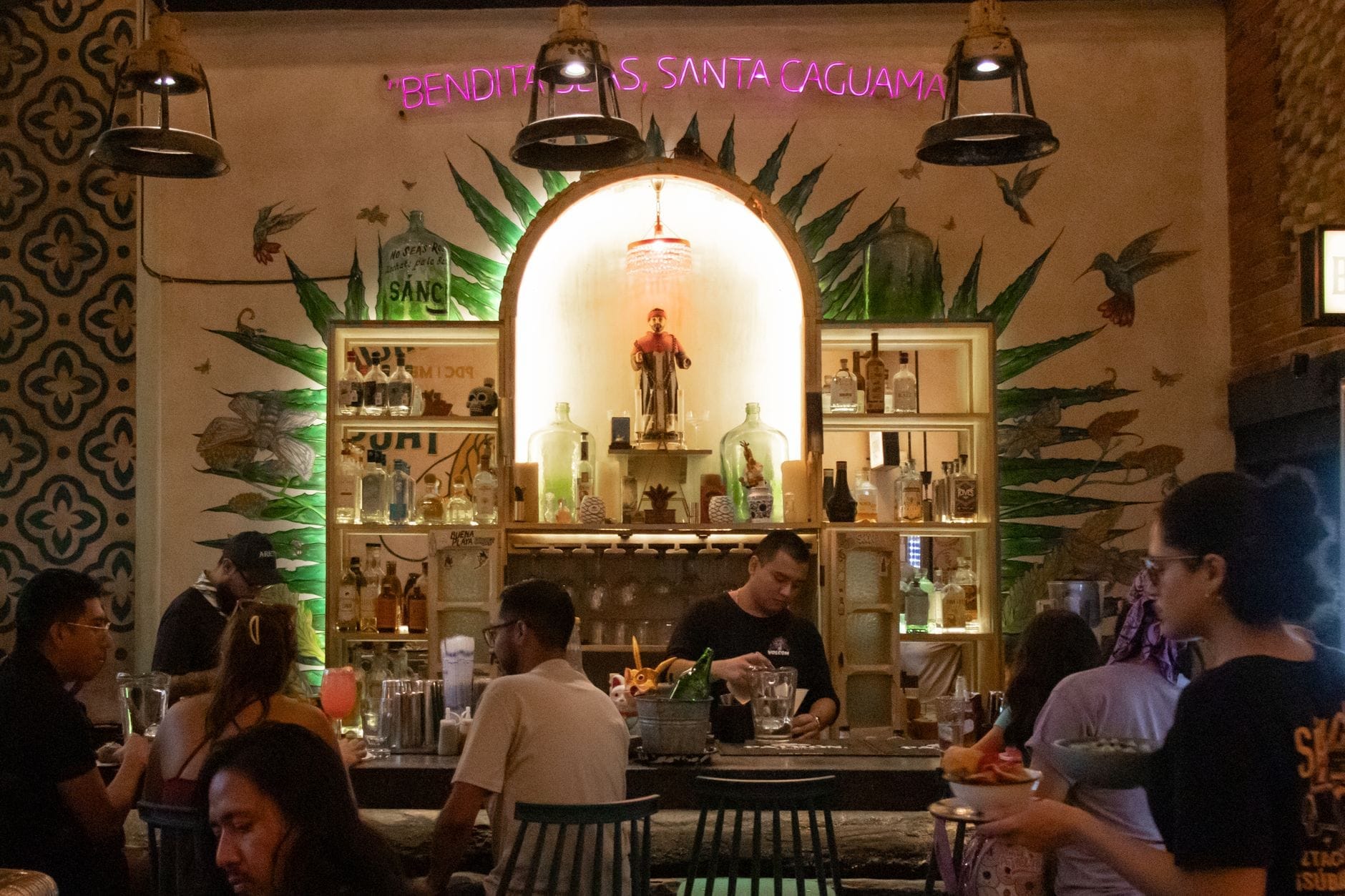 Bar de mezcal atmosférico na Cidade do México com clientes sentados no balcão, decoração de mural de agave, garrafas iluminadas por trás e bartender preparando drinques.