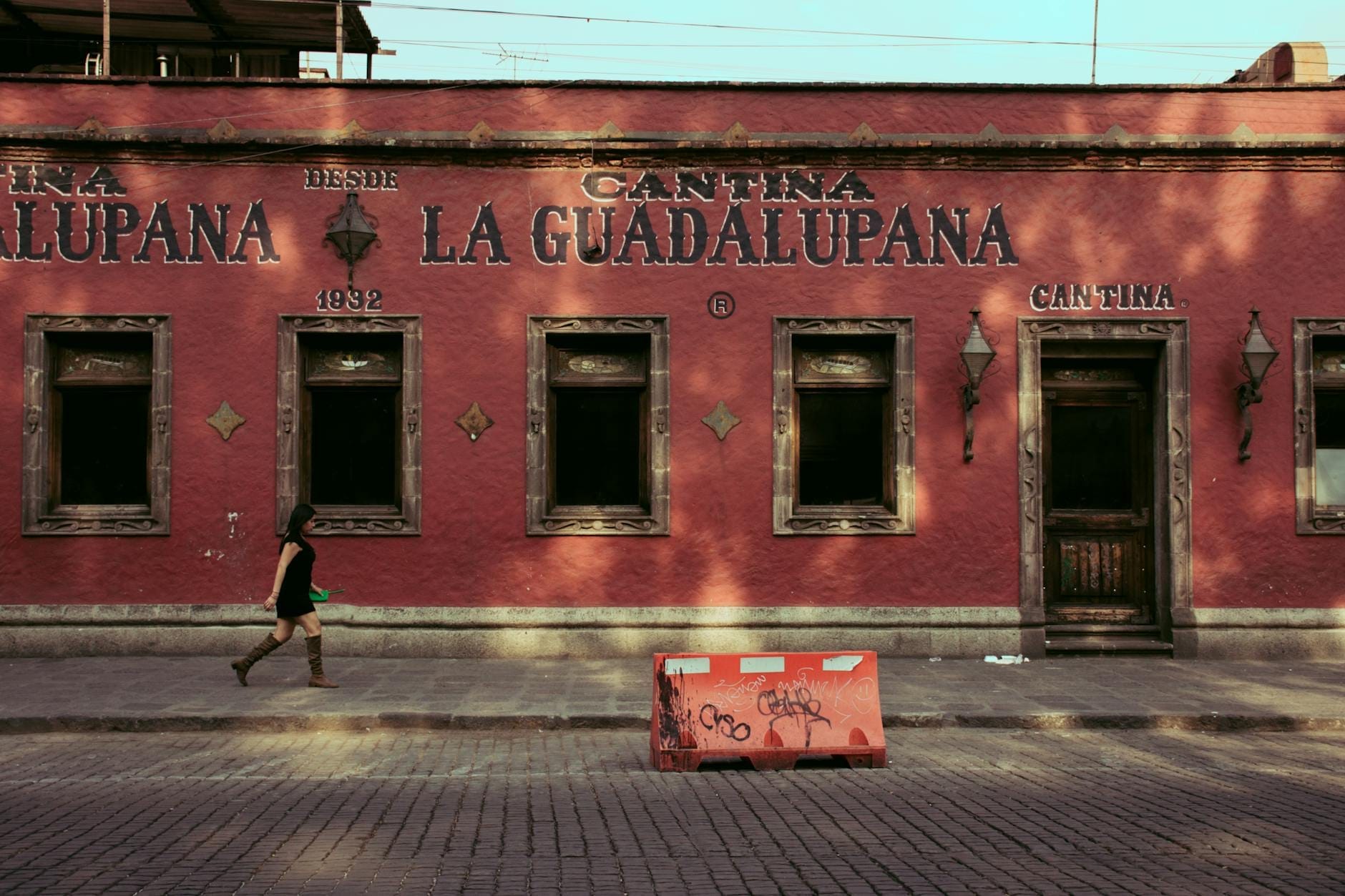 Bar cantina tradicional La Guadalupana com fachada vermelha na Cidade do México, uma mulher caminhando por uma rua de paralelepípedos.