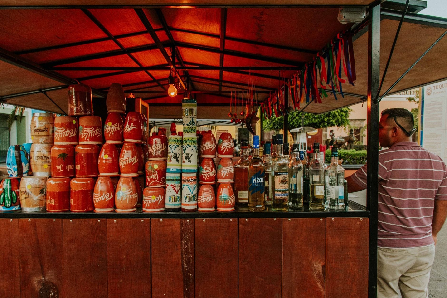 Uma barraca de mercado de madeira na Cidade do México com várias garrafas de mezcal e copos tradicionais de barro em exposição, atendida por um homem de camisa listrada.