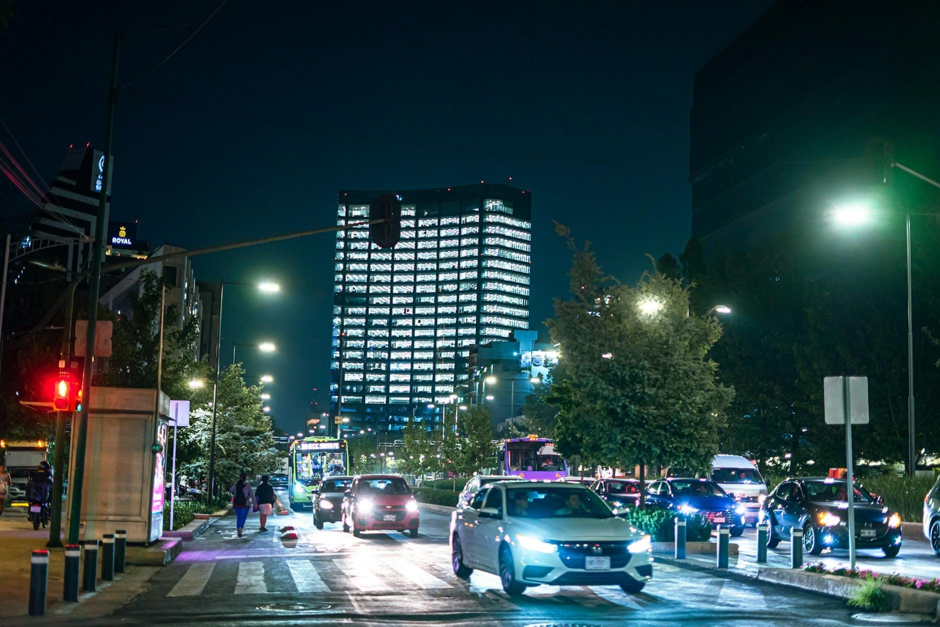 Rua da cidade do México à noite com carros, postes de luz e um alto edifício moderno iluminado ao fundo.