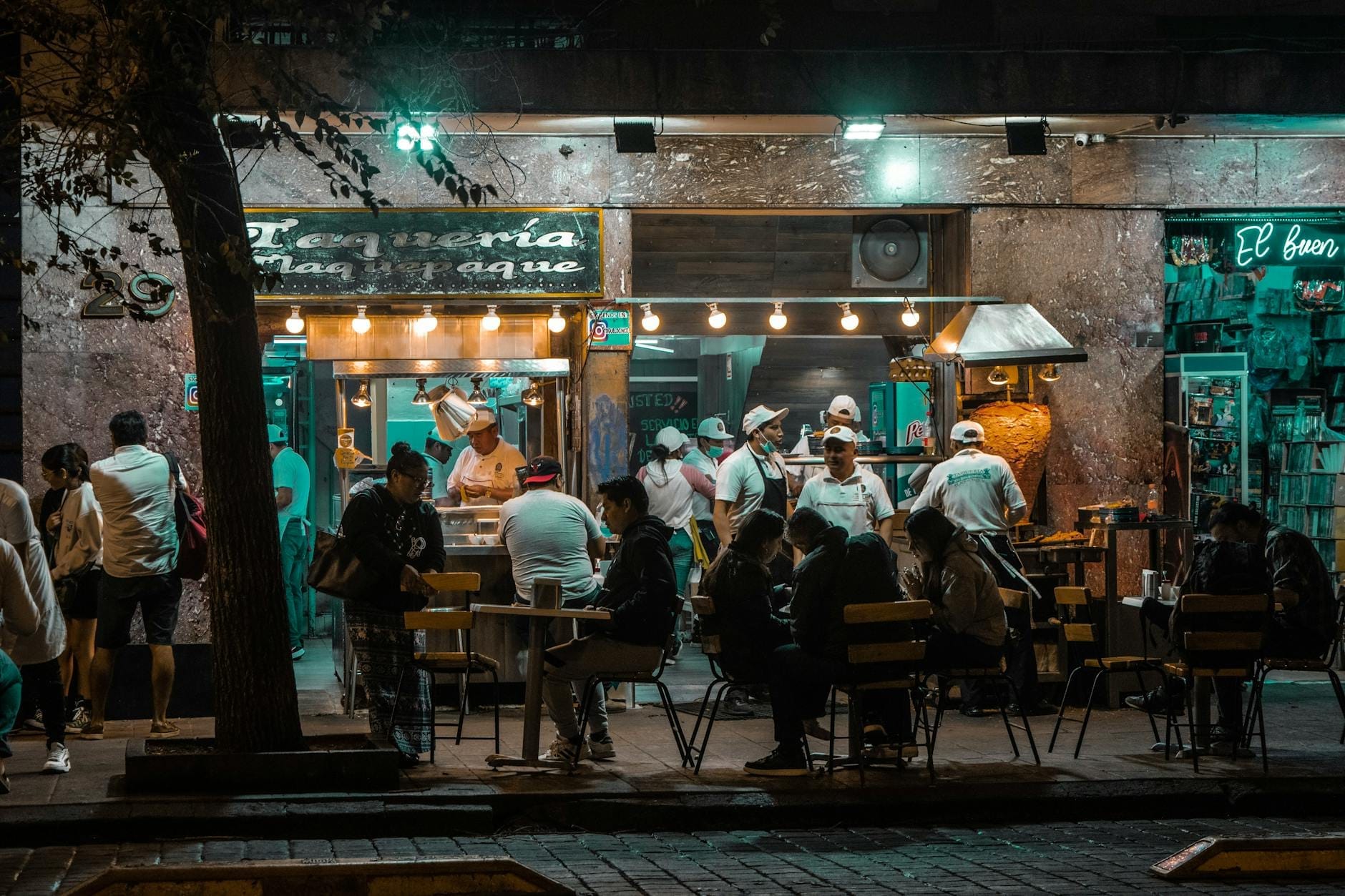 Cena noturna de uma taqueria movimentada na Cidade do México com pessoas jantando do lado de fora e cozinheiros trabalhando dentro sob luzes brilhantes.