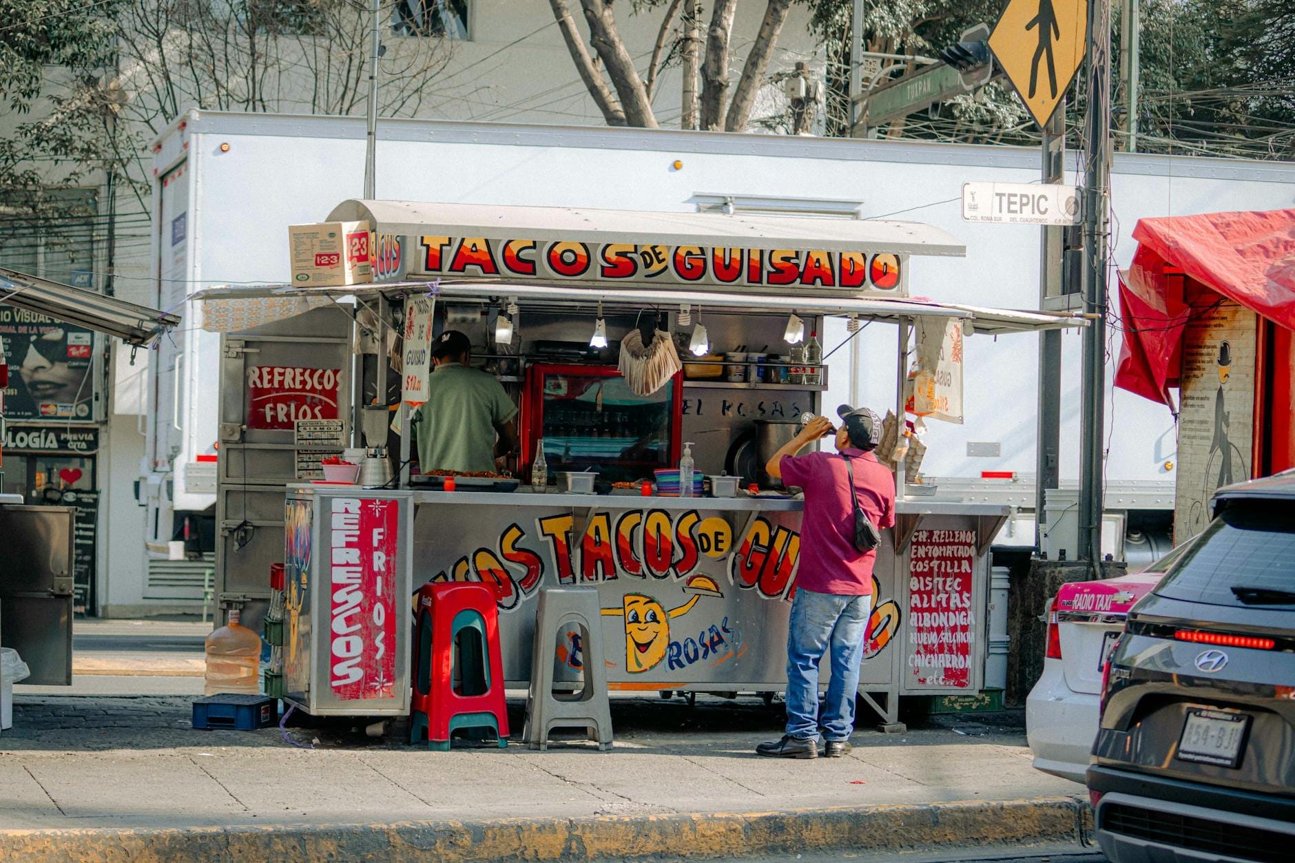 Barraca de tacos de rua com o letreiro 'Tacos de Guisado' com duas pessoas preparando comida e banquetas coloridas numa calçada da Cidade do México.