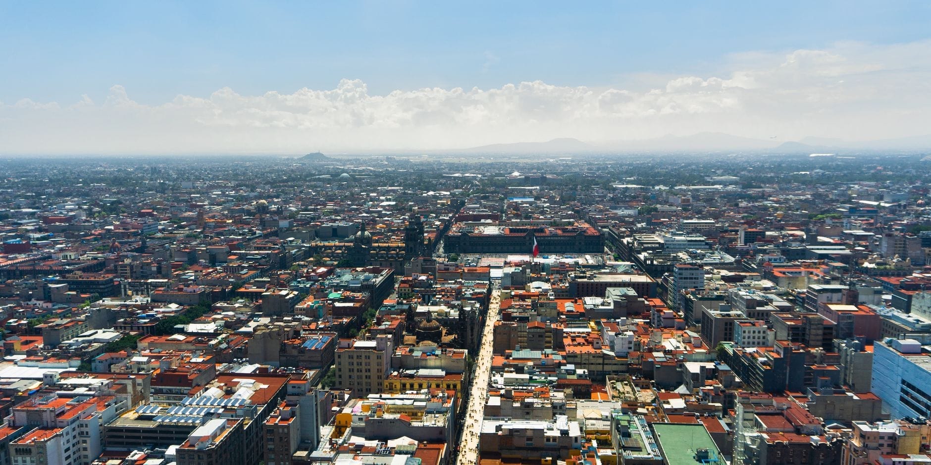 Vista aérea do centro histórico da Cidade do México com seus imensos prédios, avenidas principais e montanhas ao fundo sob um céu azul intenso, mostrando toda a grandiosidade da cidade.