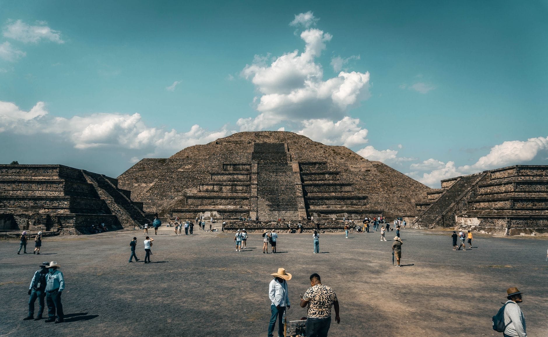 Vue de la Pyramide du Soleil de Teotihuacán avec des visiteurs traversant la place sous un ciel partiellement nuageux.