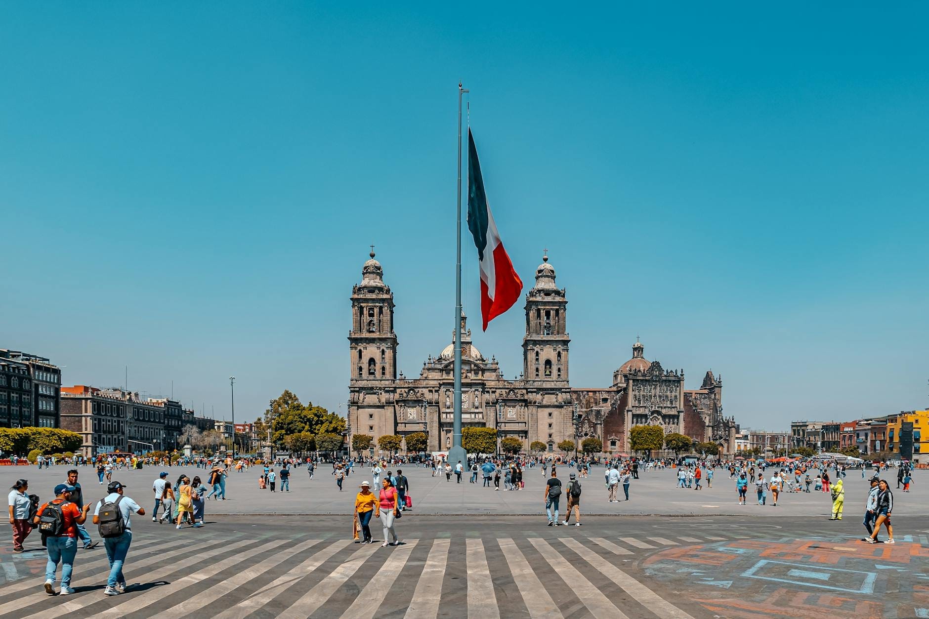 Vue large de la place avec la Cathédrale métropolitaine de Mexico et le Palais National, un grand drapeau mexicain et des personnes se promenant sur le Zócalo.