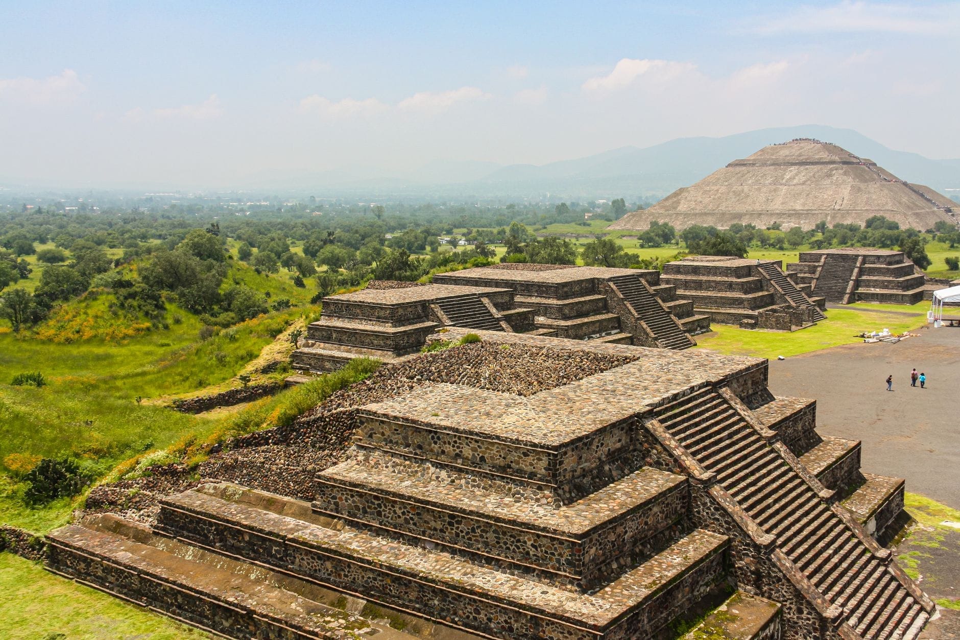 Vista ampla das pirâmides de Teotihuacán com vegetação exuberante e a Pirâmide do Sol ao fundo sob um céu luminoso, convidando à exploração.
