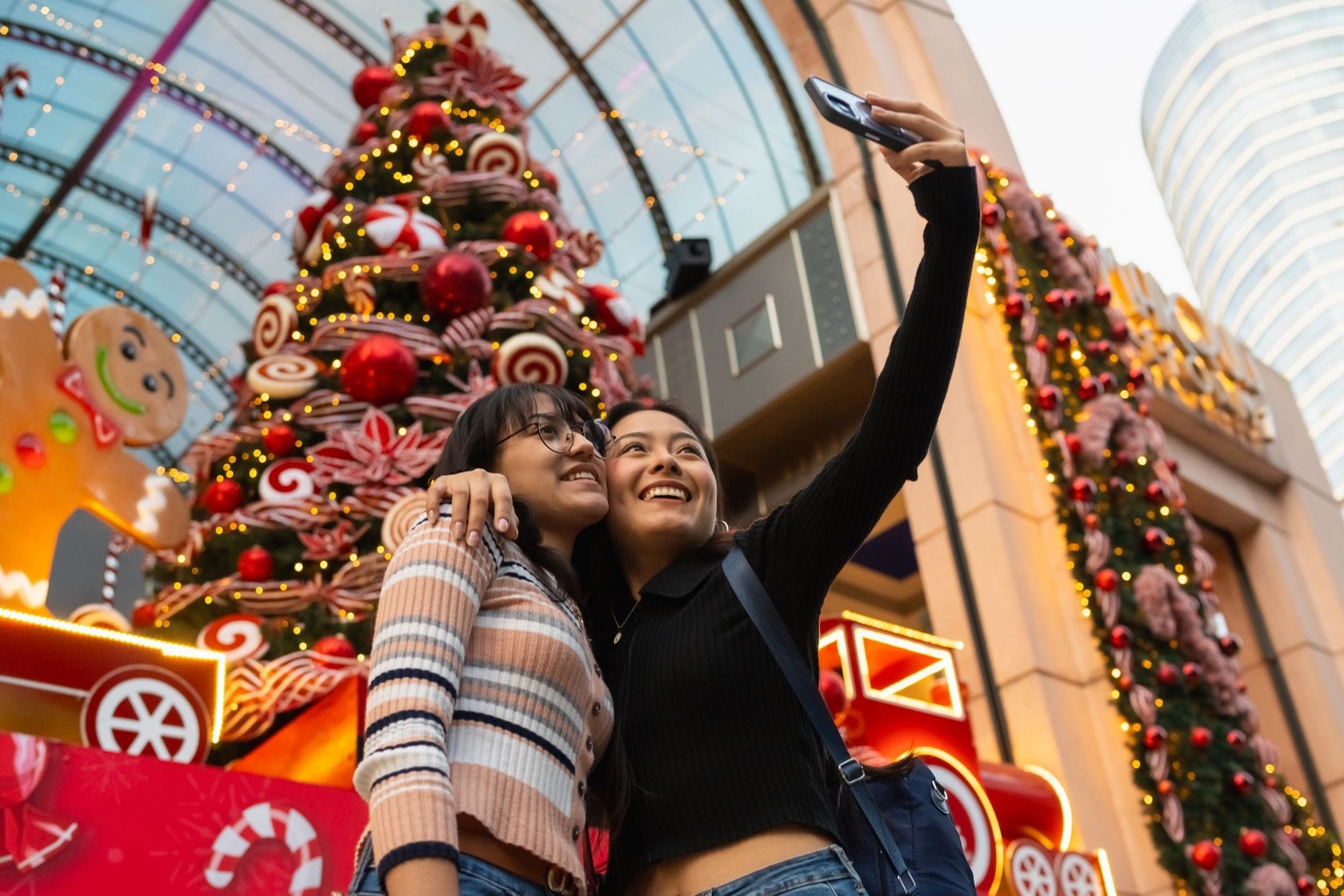 Duas mulheres tirando uma selfie em frente a um shopping center decorado com enfeites natalinos festivos e uma grande decoração de feriado.