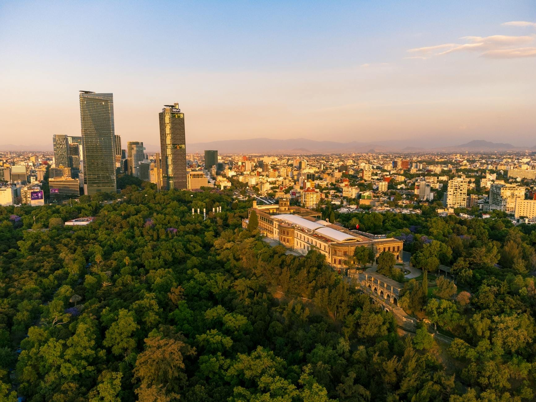 Vista aérea do Castelo de Chapultepec cercado por floresta com o horizonte da Cidade do México ao fundo durante o dia.