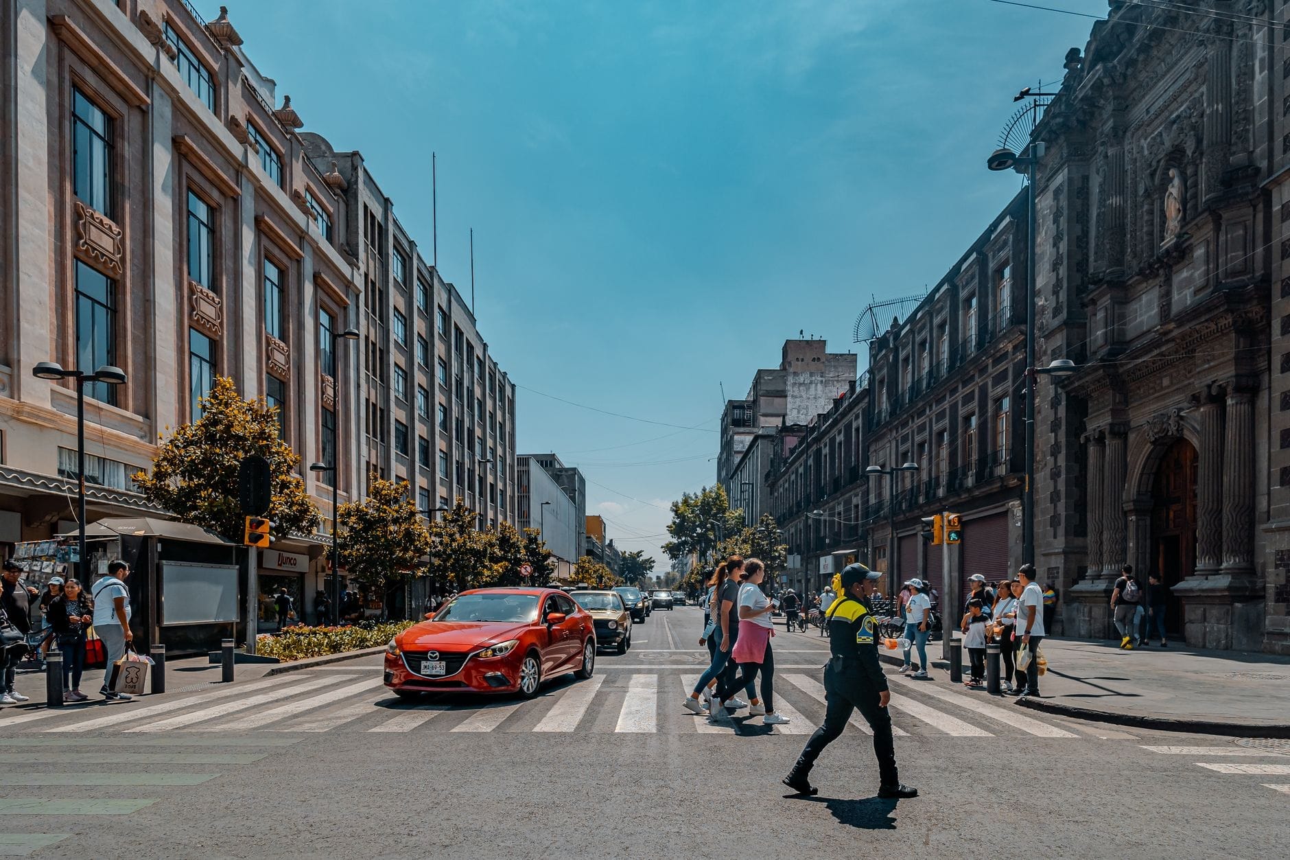 Faixa de pedestres movimentada com pedestres e um policial no centro da Cidade do México, rodeada de edifícios históricos e modernos em um dia ensolarado.