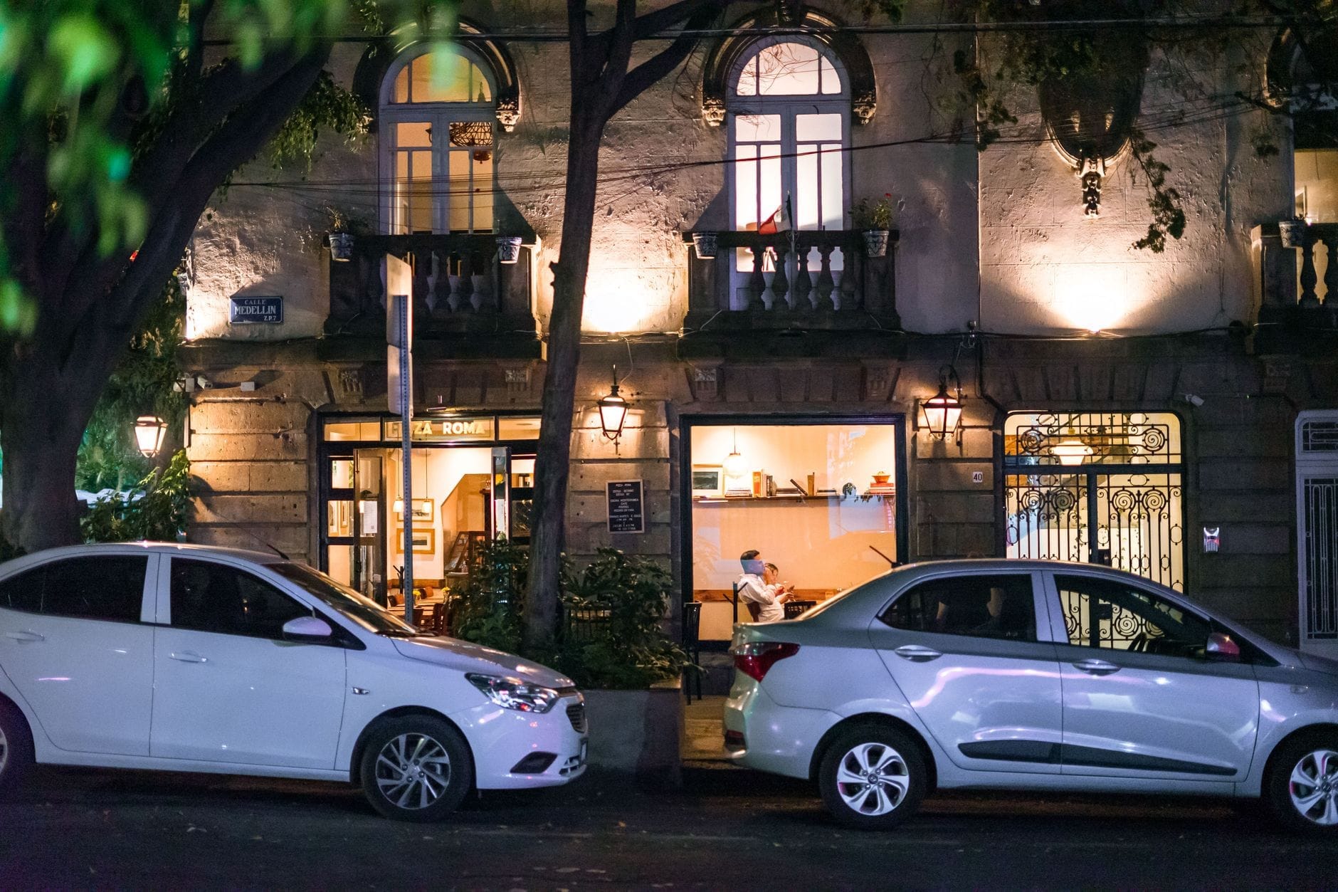 Charmosa cena noturna de uma rua bem iluminada na Cidade do México, com um elegante edifício colonial com varandas, carros estacionados e pessoas dentro de um aconchegante café.