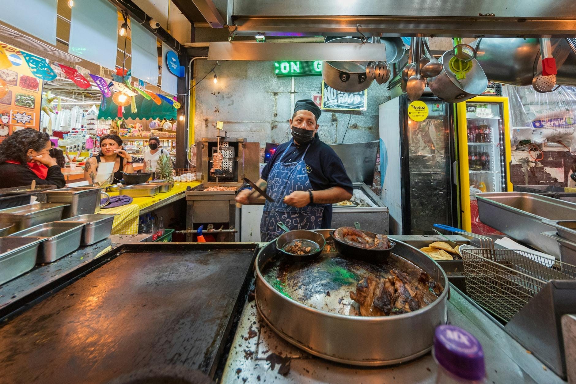 Uma barraca de comida de rua movimentada e colorida na Cidade do México com um vendedor cozinhando atrás do balcão, cercado por vários ingredientes e utensílios de cozinha.