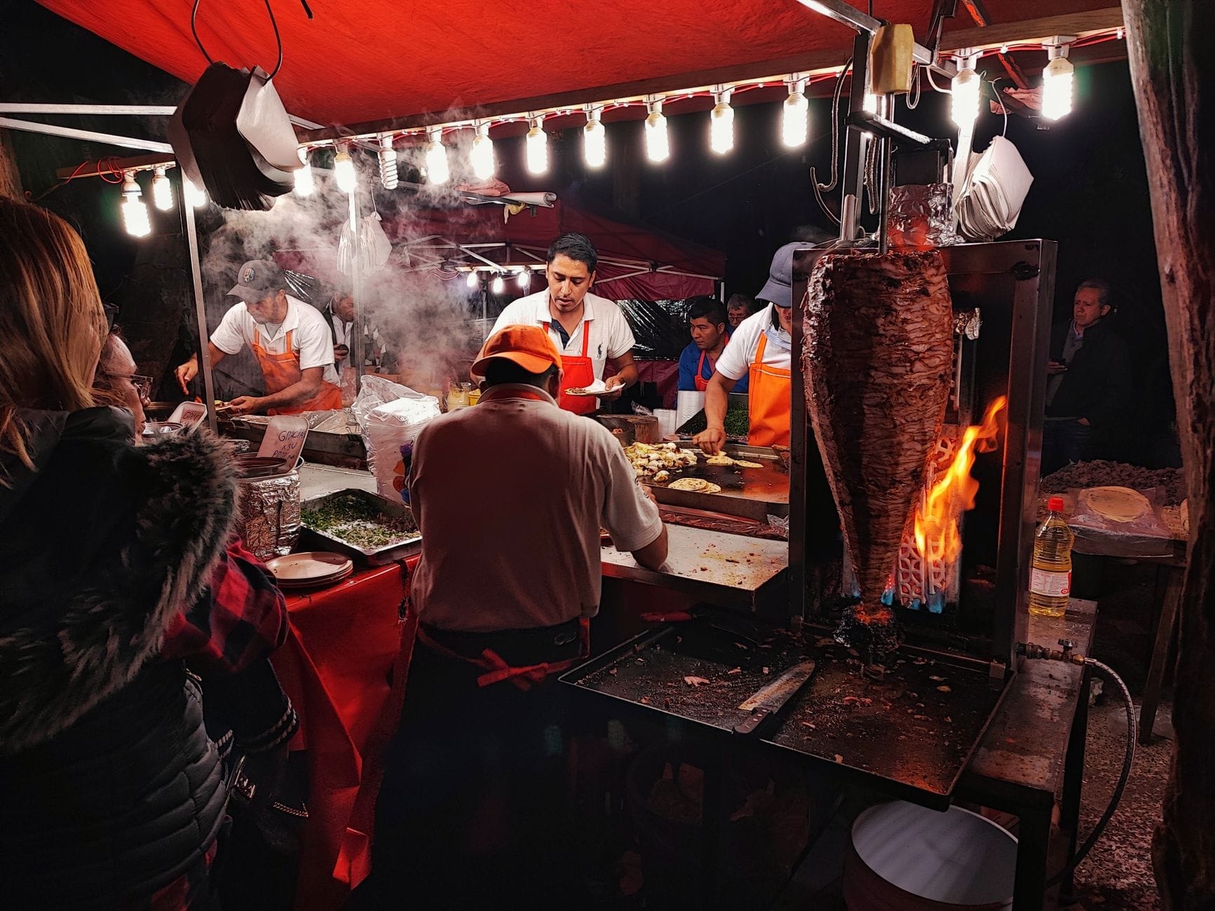 Imagem noturna de uma movimentada barraca de comida de rua na Cidade do México com trabalhadores preparando tacos al pastor, um espeto de carne e clientes aguardando.
