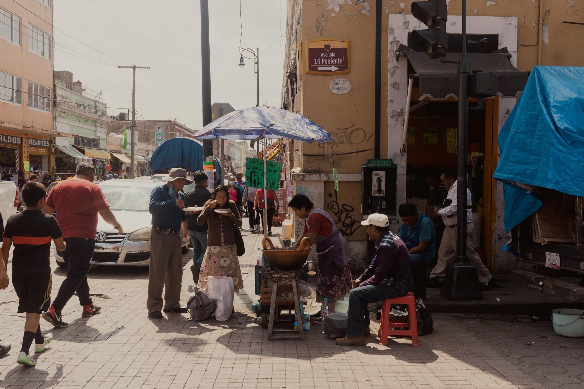 Cena de rua com pessoas reunidas ao redor de uma barraca de comida sob um guarda-sol em uma movimentada calçada da Cidade do México