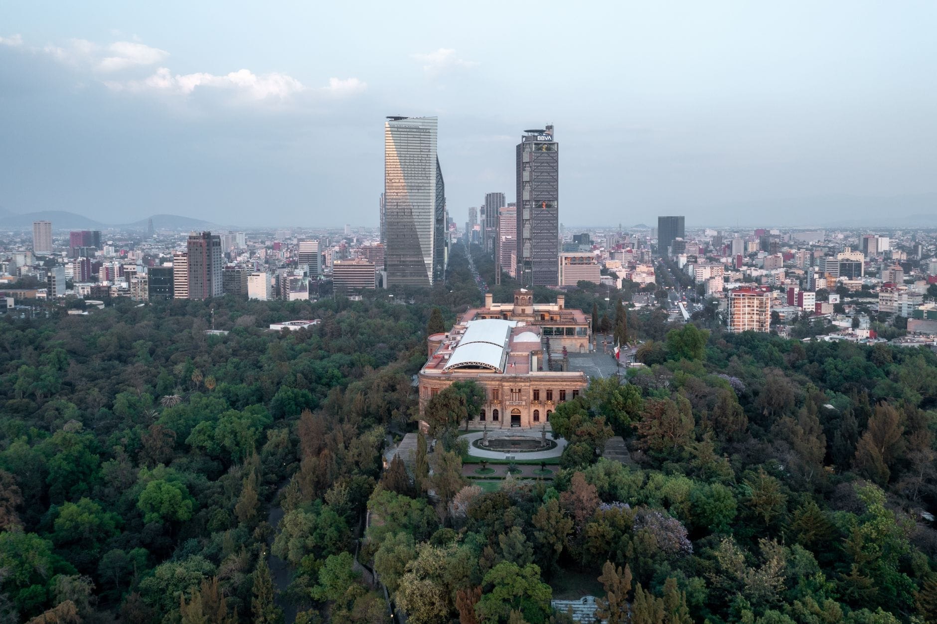 Vista aérea do Castelo de Chapultepec rodeado por áreas verdes com os arranha-céus da Cidade do México ao fundo em um dia claro.