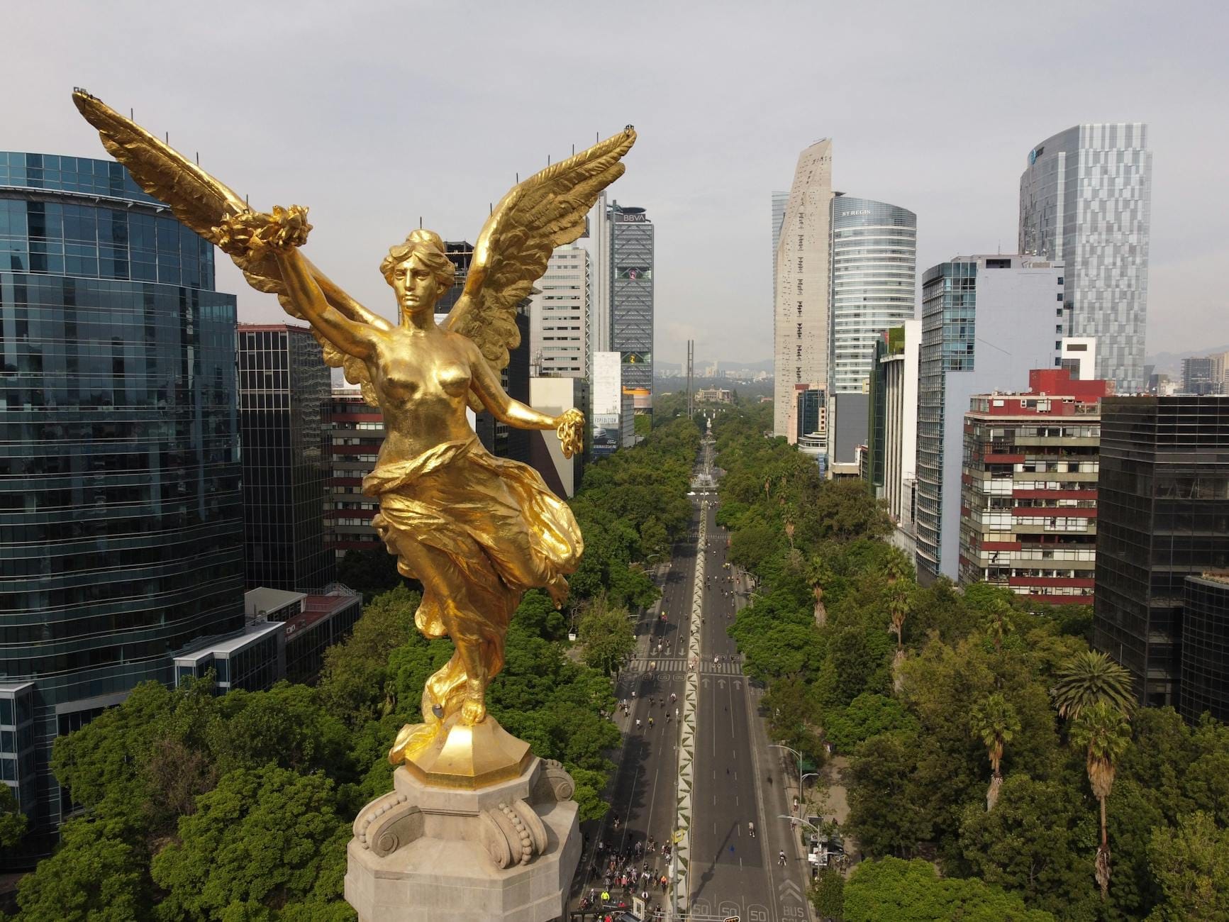 Vista aérea da dourada estátua do Ángel de la Independencia no topo de sua coluna, com o Paseo de la Reforma arborizado e o moderno horizonte urbano ao fundo.