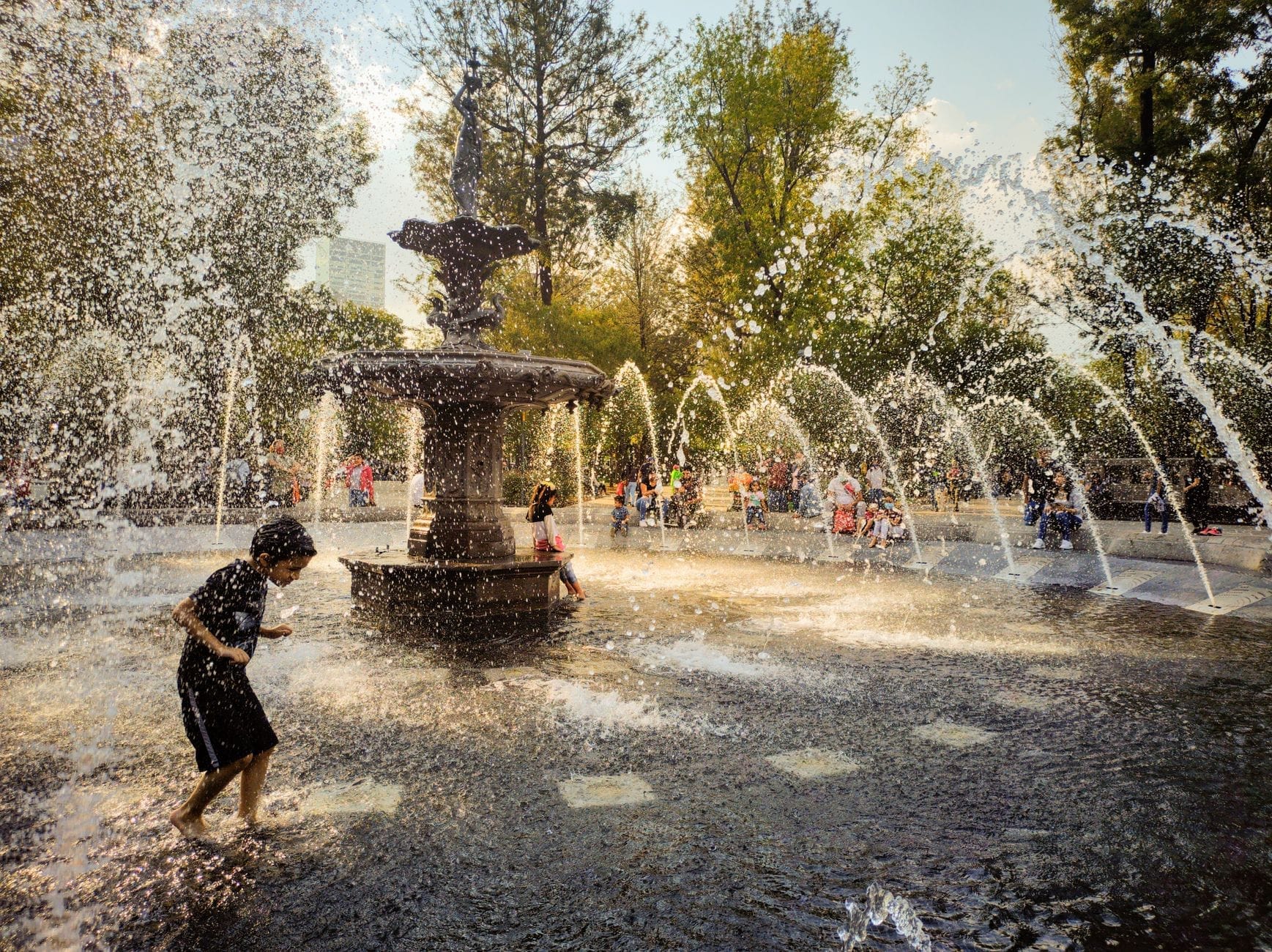 Crianças brincando e correndo entre jatos d'água de um grande chafariz em um parque arborizado e ensolarado na Cidade do México.