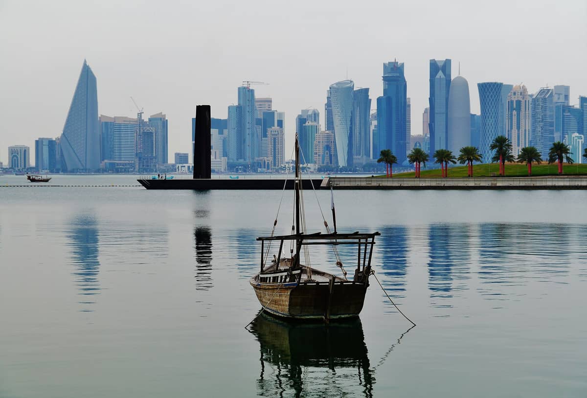 Ein traditionelles hölzernes Dhau-Boot treibt auf ruhigem Wasser, im Vordergrund Palmen und die grünen Rasenflächen des MIA Parks, im Hintergrund die moderne Skyline von Dohas West Bay.