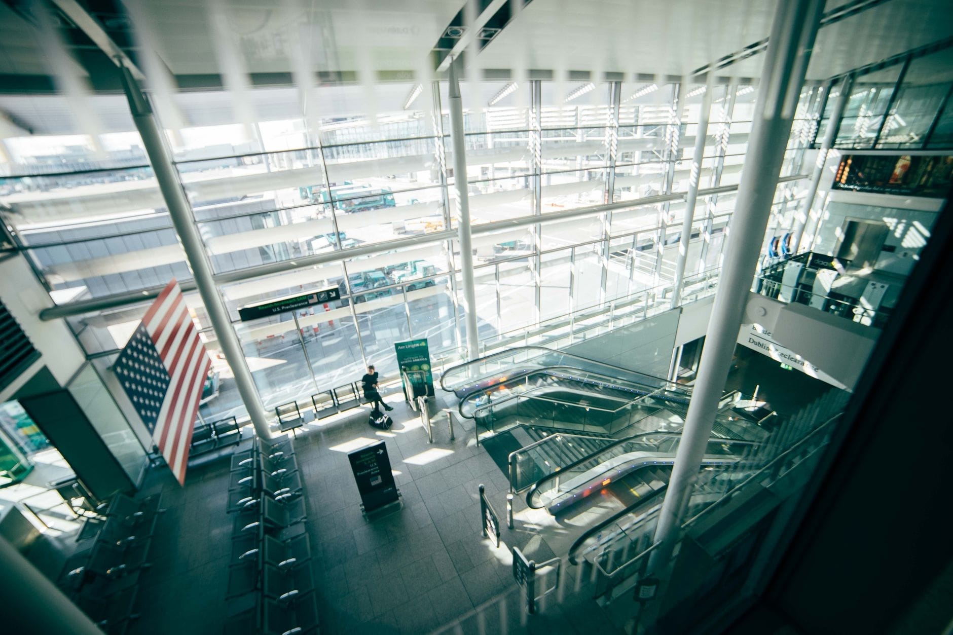 Interior moderno e iluminado de um terminal de aeroporto com grandes janelas, escadas rolantes e uma bandeira americana em destaque, mostrando uma ampla área de chegadas durante o dia.
