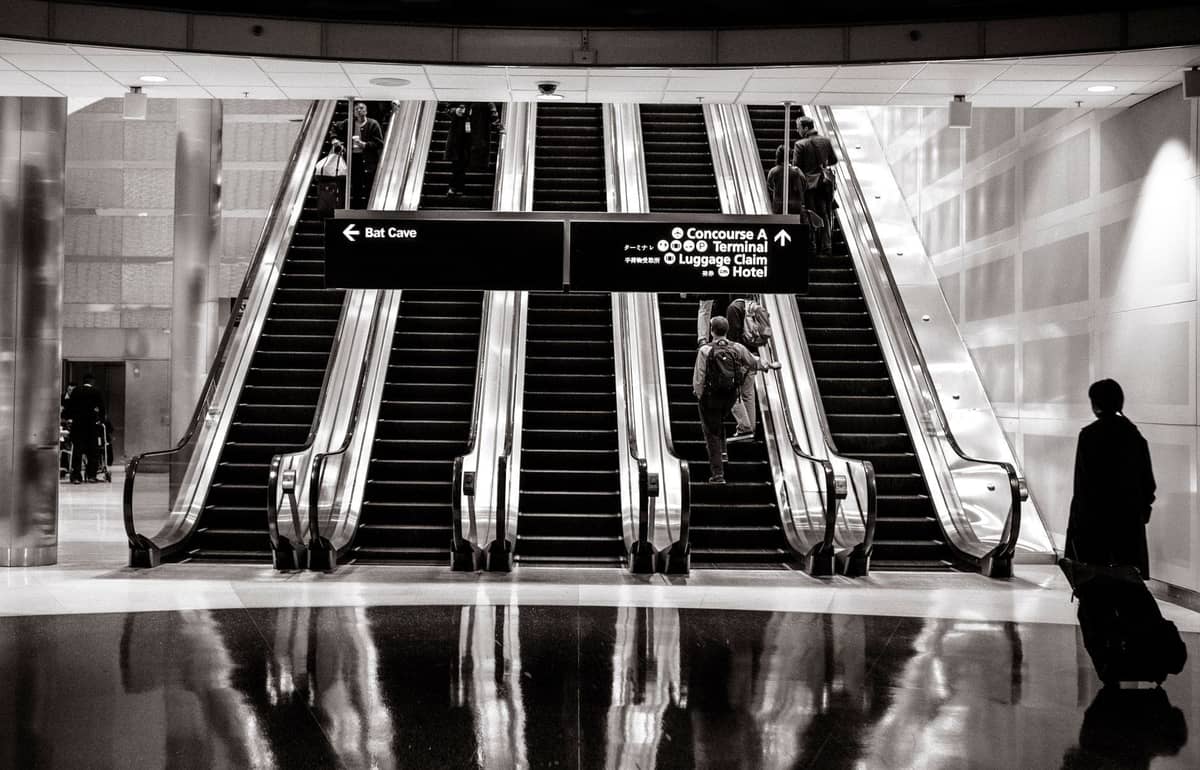 Travelers with suitcases riding escalators under airport signage showing directions to concourses, terminals, luggage claim, and hotel.
