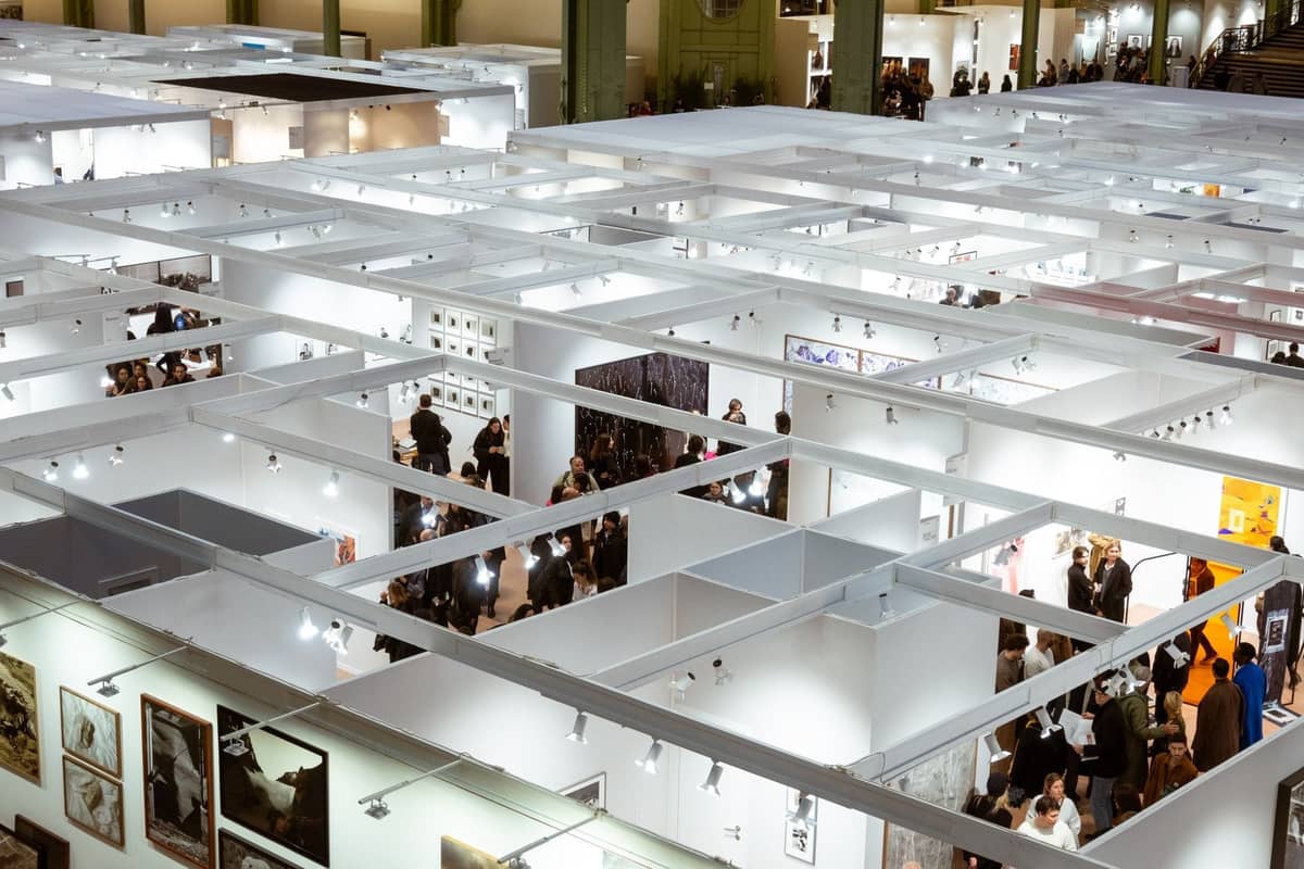 Overhead view of a bustling contemporary art exhibition with people walking through white-walled booths displaying modern artworks.