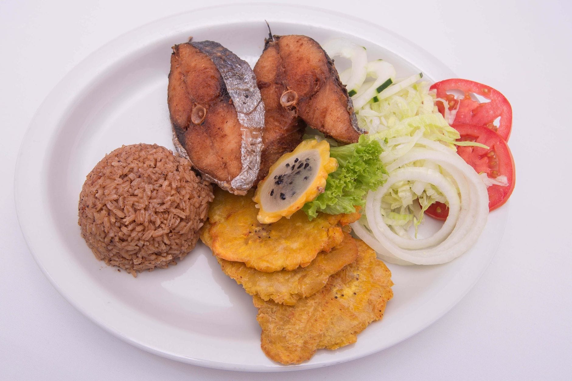 Um prato com comida cubana clássica: peixe frito, tostones (bananas verdes fritas), arroz com feijão, alface, cebola e tomate, servido sobre fundo branco.