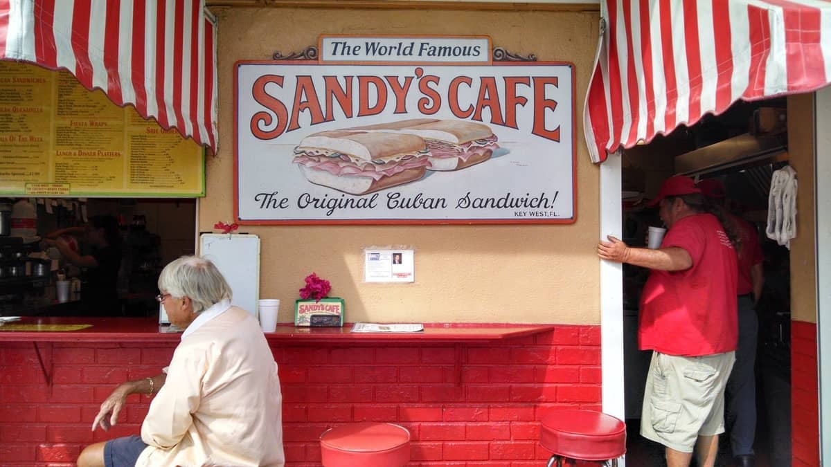 Streetfront counter of Sandy's Cafe with a sign advertising Cuban sandwiches and people sitting and ordering food.