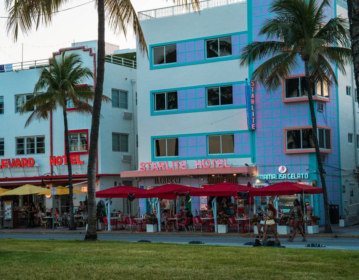 Outdoor dining scene at Miami's iconic pastel art deco hotels, with patio tables, umbrellas, and palm trees, suggesting a warm climate and relaxed atmosphere.