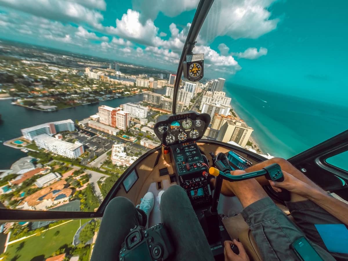 A couple in a helicopter cockpit flying over Miami Beach, with the city, coastline, and turquoise ocean visible below.