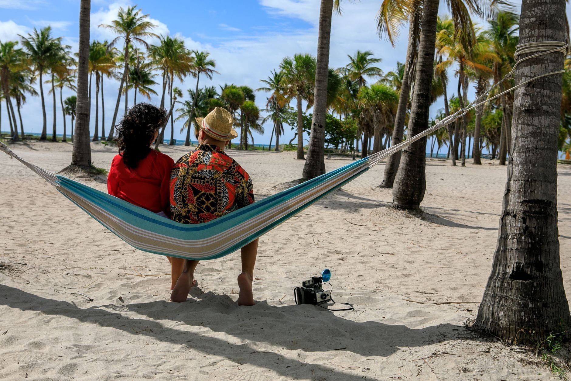 Casal relaxando em uma rede esticada entre palmeiras na areia de uma praia em Miami, com palmeiras exuberantes e céu azul ao fundo.