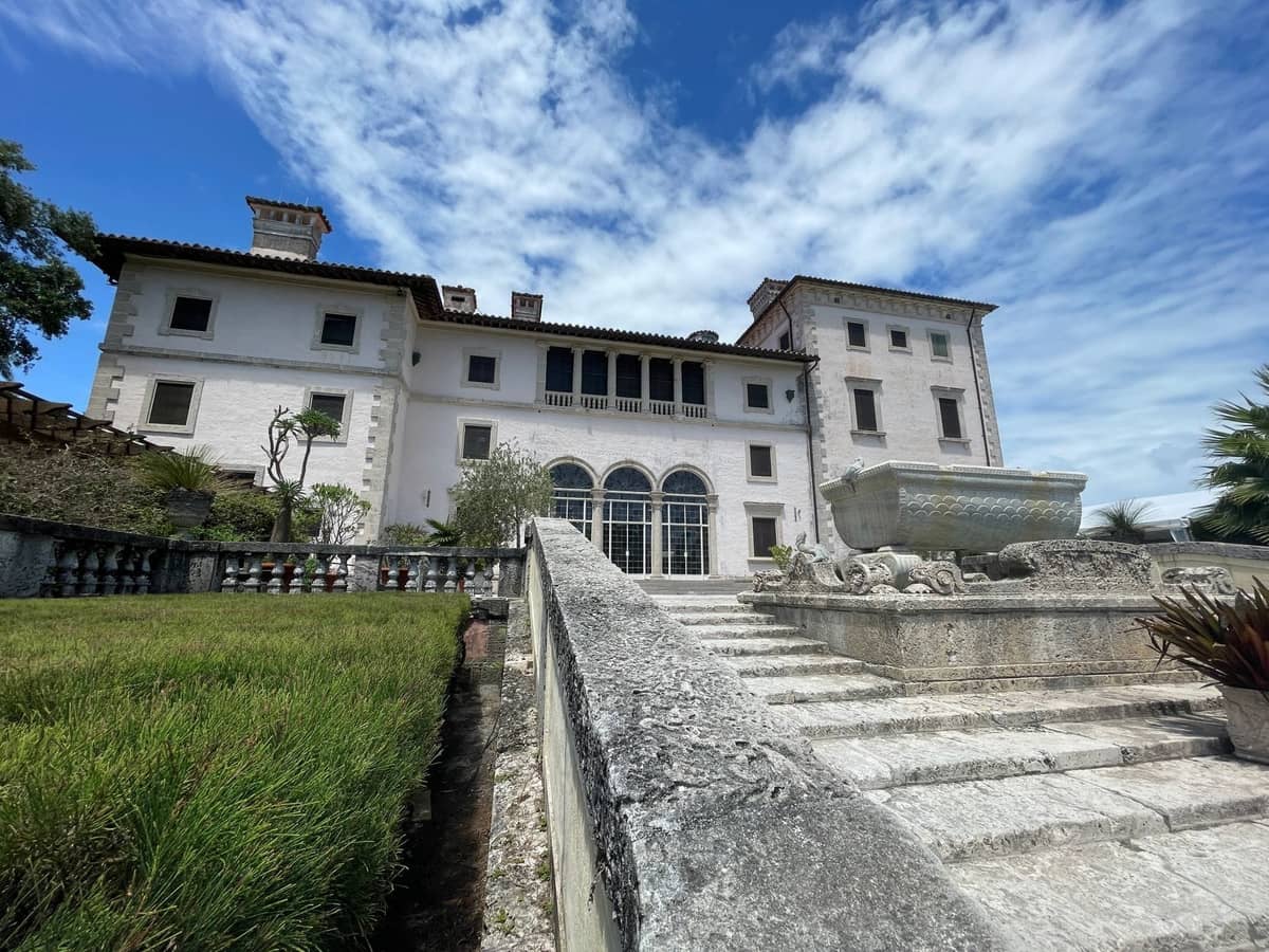 Stone staircase leads up to the façade of an Italianate villa with arched windows, reflecting a bright blue sky and scattered clouds.