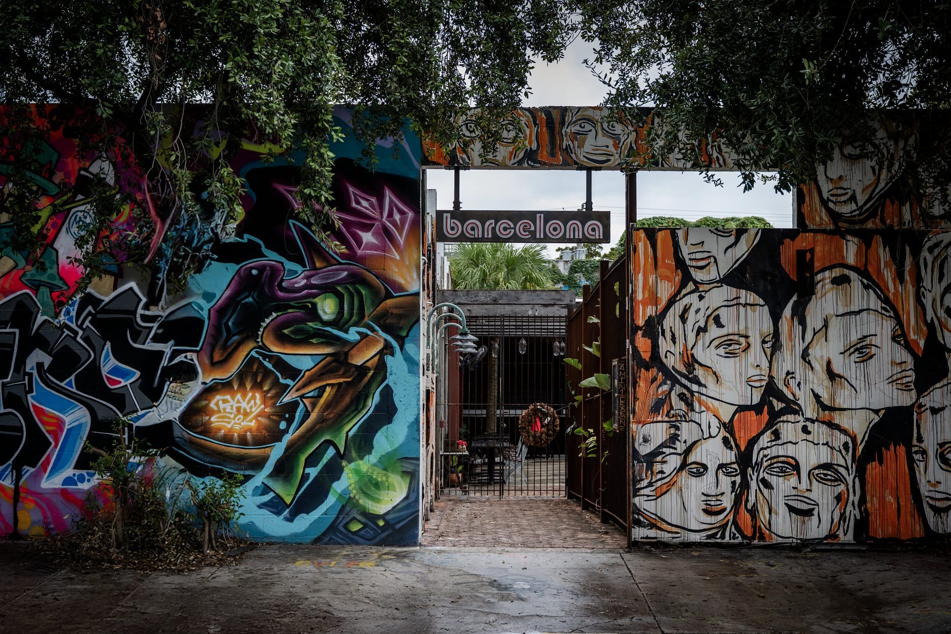 Entrada de um pátio escondido em Miami ladeado por grafites e murais coloridos, com árvores frondosas formando um arco sobre a passagem e mesas visíveis lá dentro.