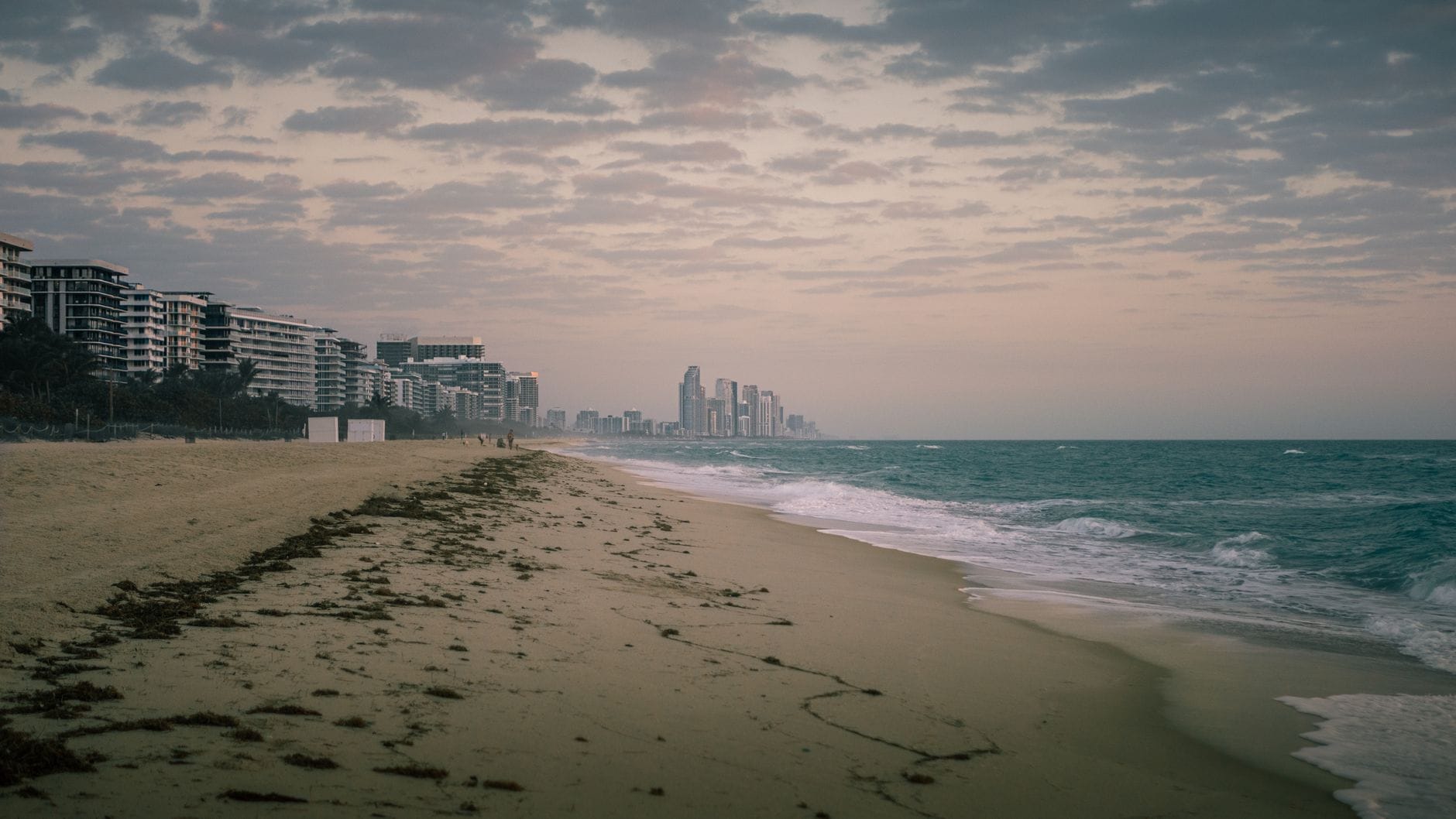 Vista ampla da orla de Miami Beach no inverno, com modernos edifícios altos, céu nublado e ondas suaves ao longo da costa arenosa.