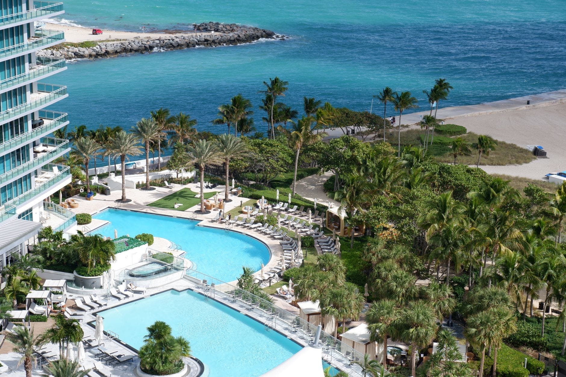 Vista aérea de um resort de luxo à beira-mar em Miami com várias piscinas, palmeiras, cabanas e acesso direto à praia de areia branca e água turquesa.