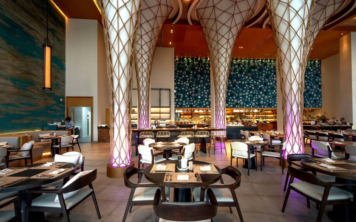 Upscale, spacious restaurant interior with modern architecture, elegant columns, and stylish lighting, featuring well-set tables and a vibrant bar in the background.