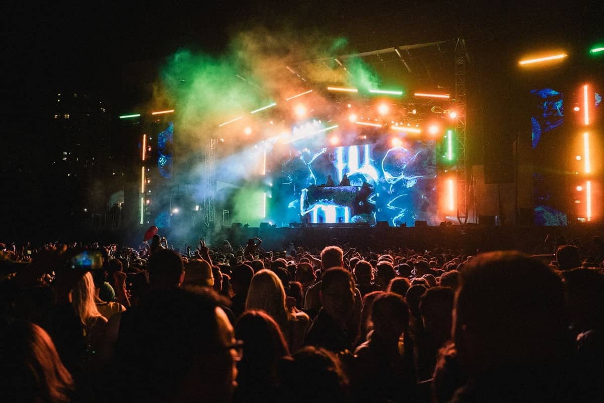 Large crowd at a nighttime outdoor electronic music festival with vibrant stage lights and smoke effects