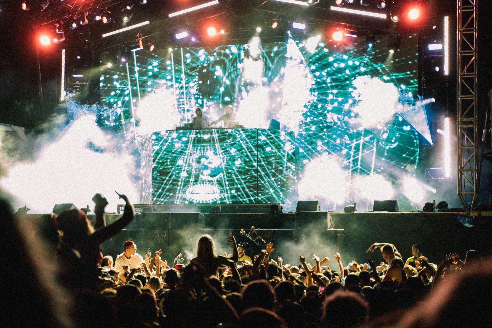Grande multidão em um festival noturno em frente a um palco iluminado com visuais eletrônicos e efeitos de fumaça, transmitindo a energia intensa de um evento de música ao ar livre.