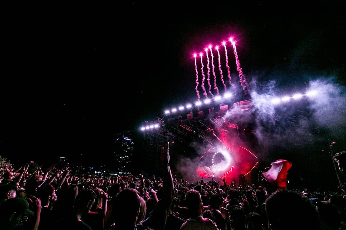 Nighttime music festival with large crowd, raised hands, bright stage lighting, fireworks, city buildings in background and EDM atmosphere.