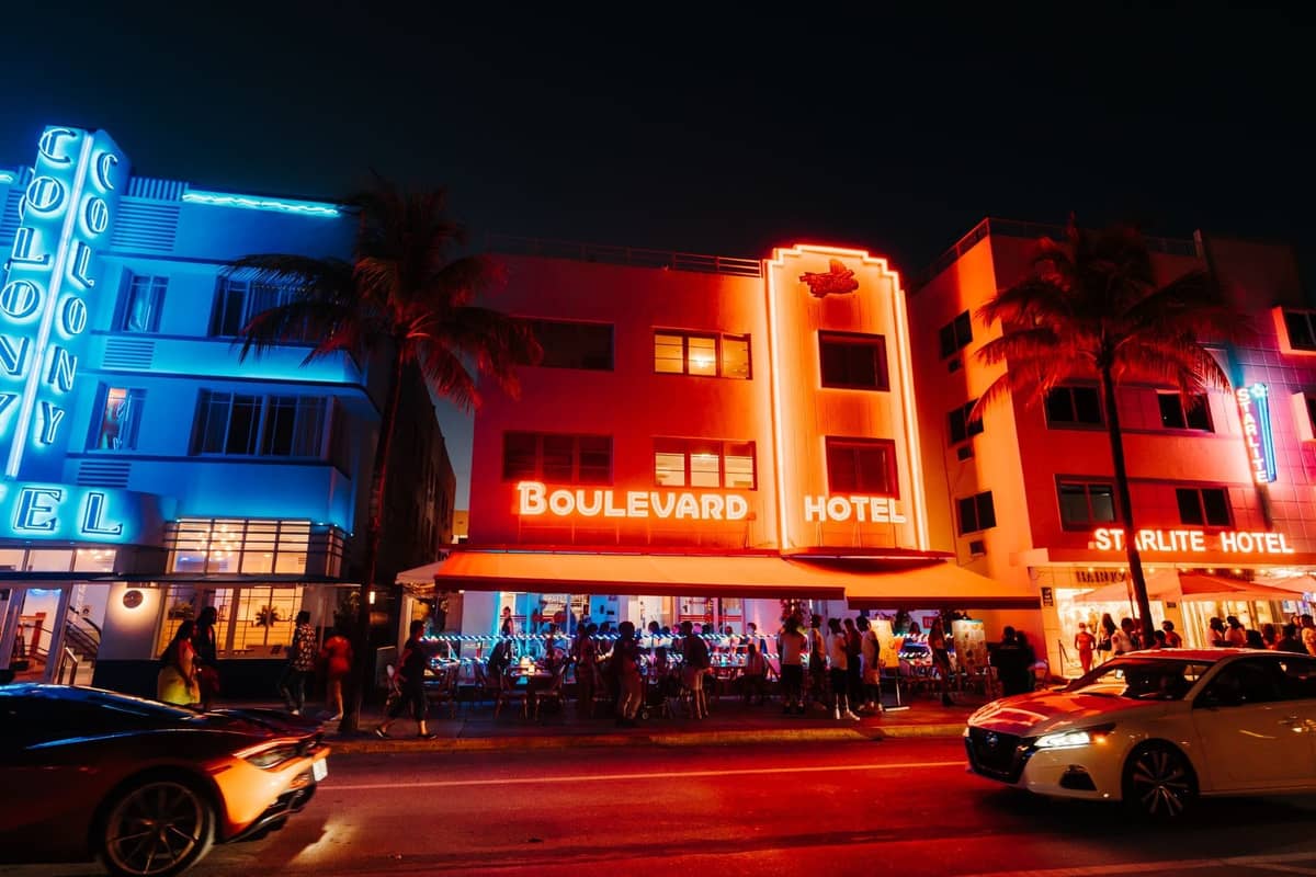 Iconic neon-lit hotels and crowded sidewalk on Ocean Drive in Miami Beach at night, bustling with nightlife activity.
