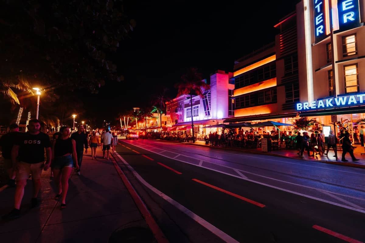 Miami's Ocean Drive at night, crowds walking by colorful neon-lit Art Deco buildings and busy outdoor bars and restaurants.