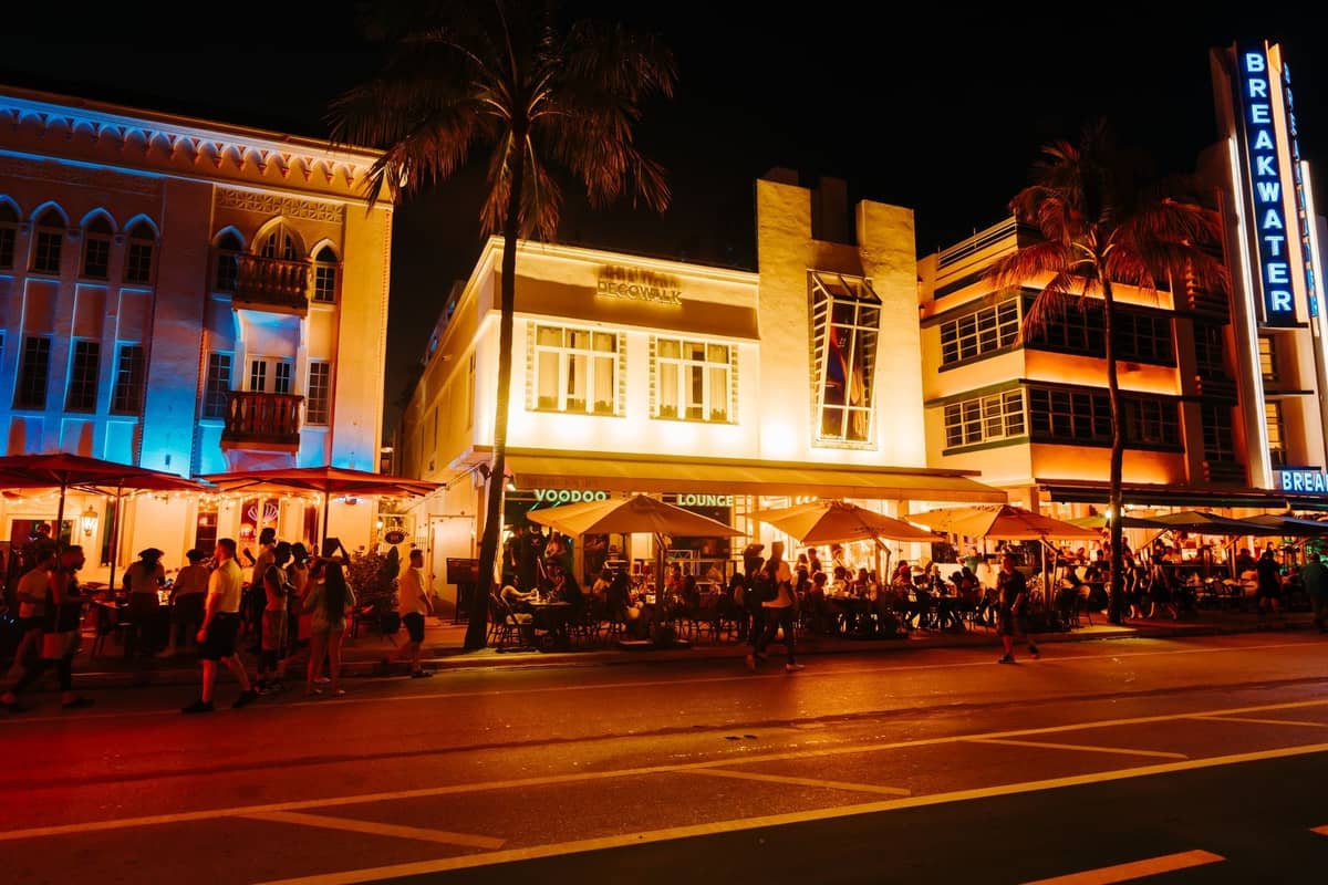Night scene of Miami's Ocean Drive with lively crowds dining outdoors at vibrant restaurants under neon lights.
