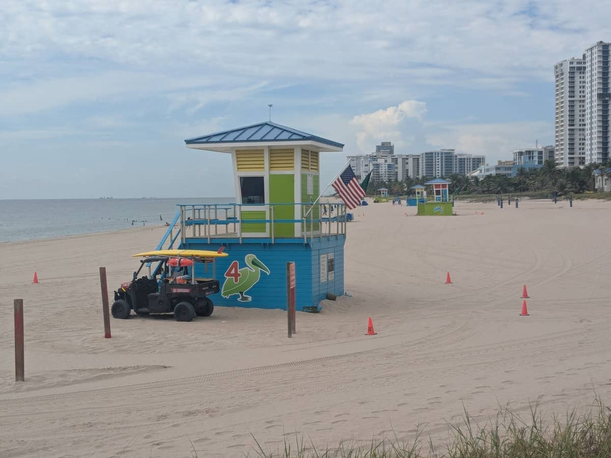 Miami beach scene with colorful lifeguard tower, nearby high-rise hotels, and a mostly empty sandy shore on a sunny day.