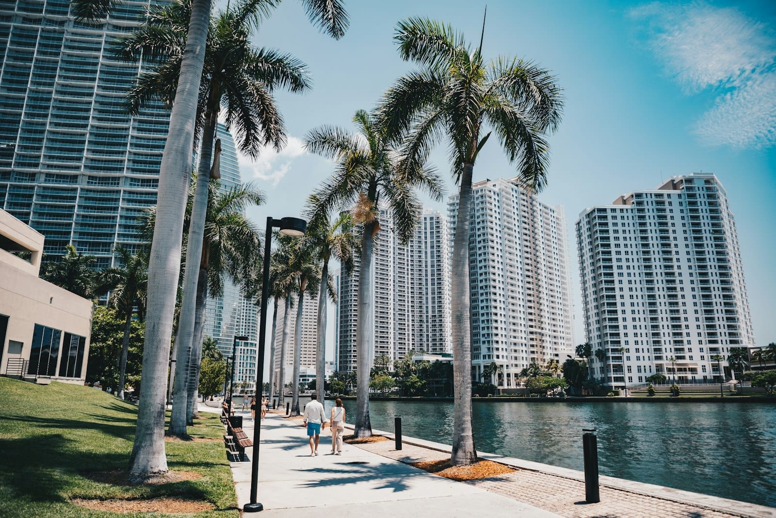 Persone che passeggiano lungo il Miami Riverwalk fiancheggiato da palme, con grattacieli moderni e il fiume visibile in una giornata di sole.
