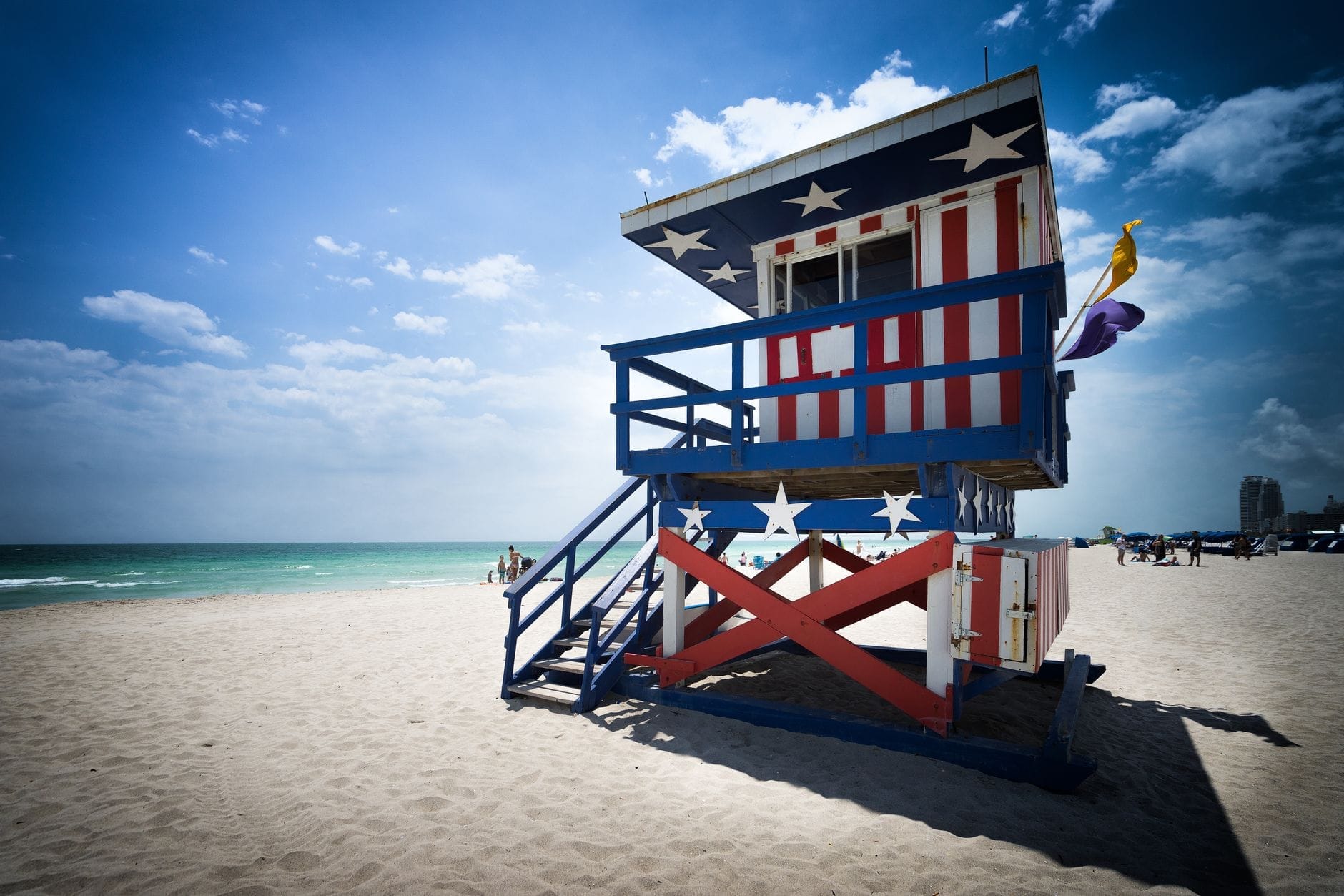 Torre de salva-vidas com listras vermelhas, brancas e azuis na Miami Beach, com céu limpo, areia e mar ao fundo — ícone local que transmite segurança e o charme da cidade.
