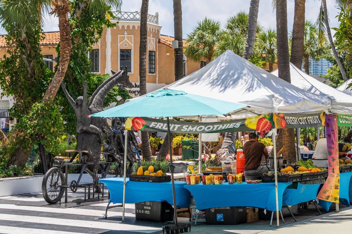 Outdoor market scene in Miami with fruit stands under tents, palm trees, and Mediterranean-style buildings in the background.