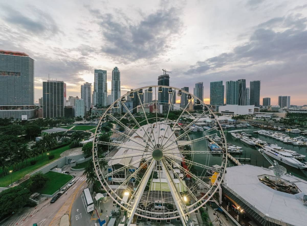View of Bayside Marketplace, Ferris wheel, Port Miami, and city skyline at sunset, with road and marina in foreground.