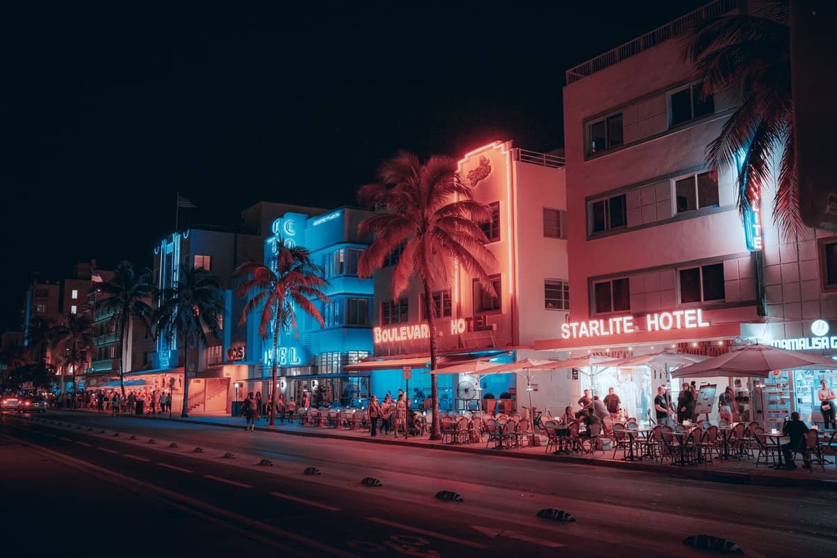 Nighttime view of Miami’s Ocean Drive with neon-lit Art Deco hotels, palm trees, outdoor diners, and a lively street scene, evoking iconic Miami Vice atmosphere.